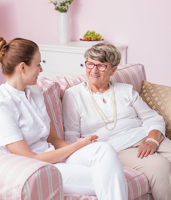 Caregiver and elderly woman smiling and talking while seated together on a couch.